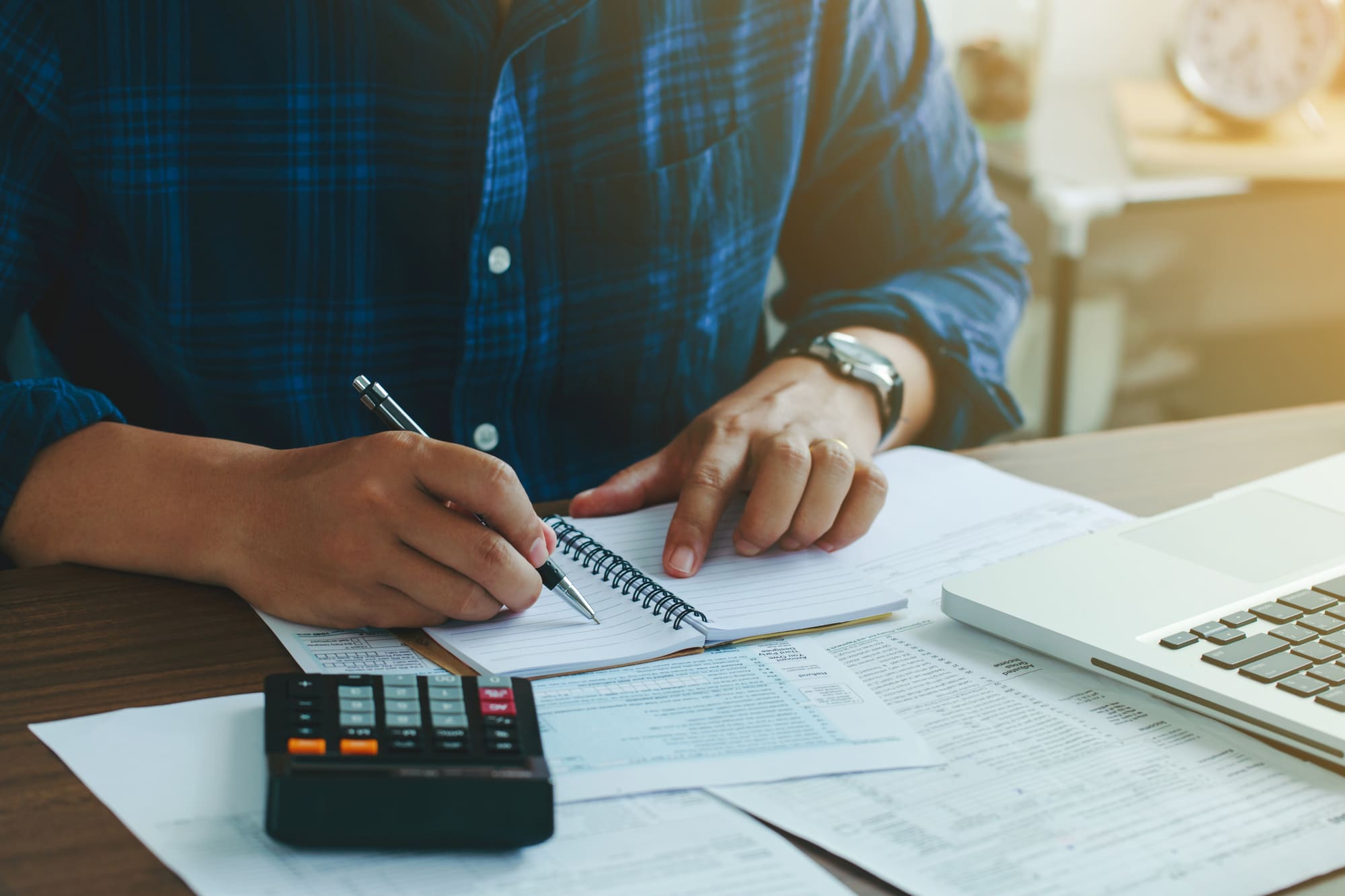 Person in blue plaid shirt writing in spiral notebook at desk with calculator and financial documents